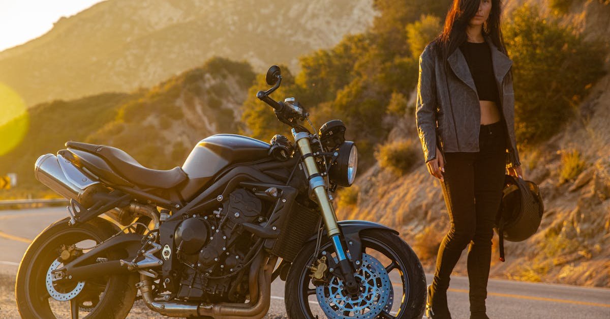 Woman standing beside motorcycle on a sunlit scenic roadside, holding a helmet.